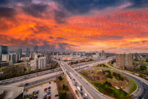Urban cityscape showing a busy highway interchange with cars driving under a dramatic orange and red sunset sky, highlighting infrastructure and modern living